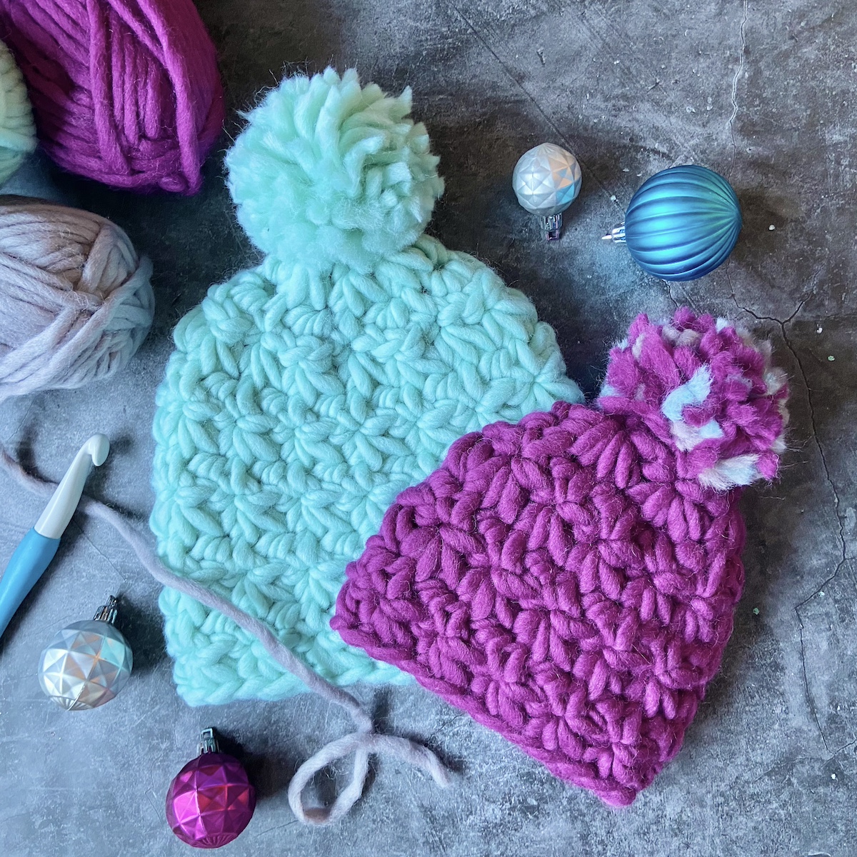 Two crocheted hats made with chunky yarn sitting on the floor with Christmas ornaments and yarn