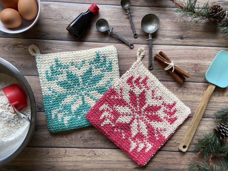 one pink and one blue snowflake tapestry crochet potholders laying on wood paneling surrounded by baking supplies