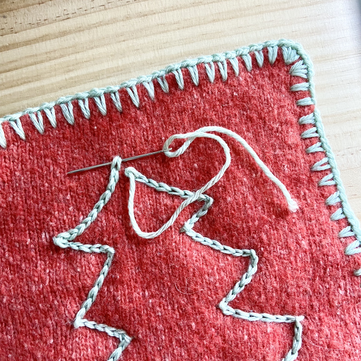 a stitched outline of a blue yarn Christmas tree on red sweater material with a sewing needle embedded in the stitches