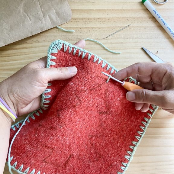 hands holding a crochet hook and upcycled sweater potholder beginning to crochet a Christmas tree motif in the center