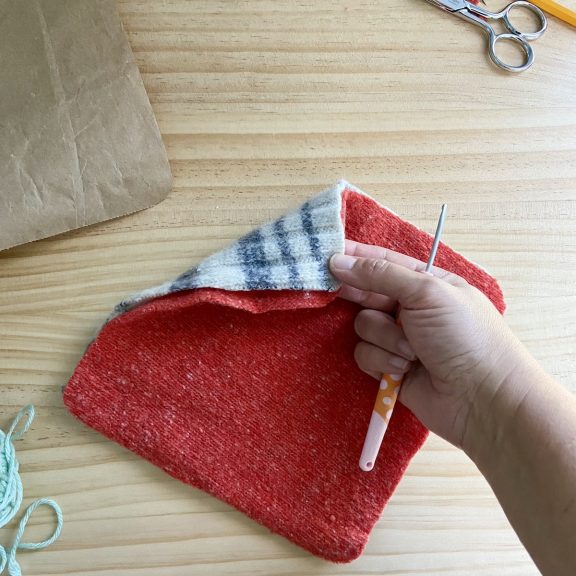 a hand holding a crochet hook and pinching two squares of sweater material held together