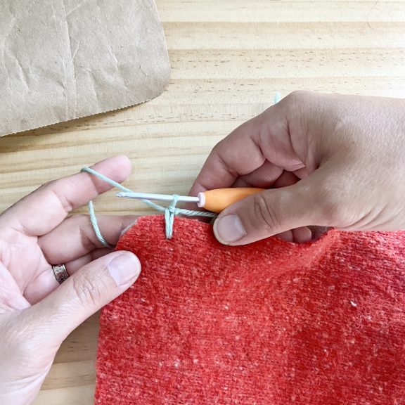 two hands holding a crochet hook and red sweater fabric creating a knot in the first yarn over