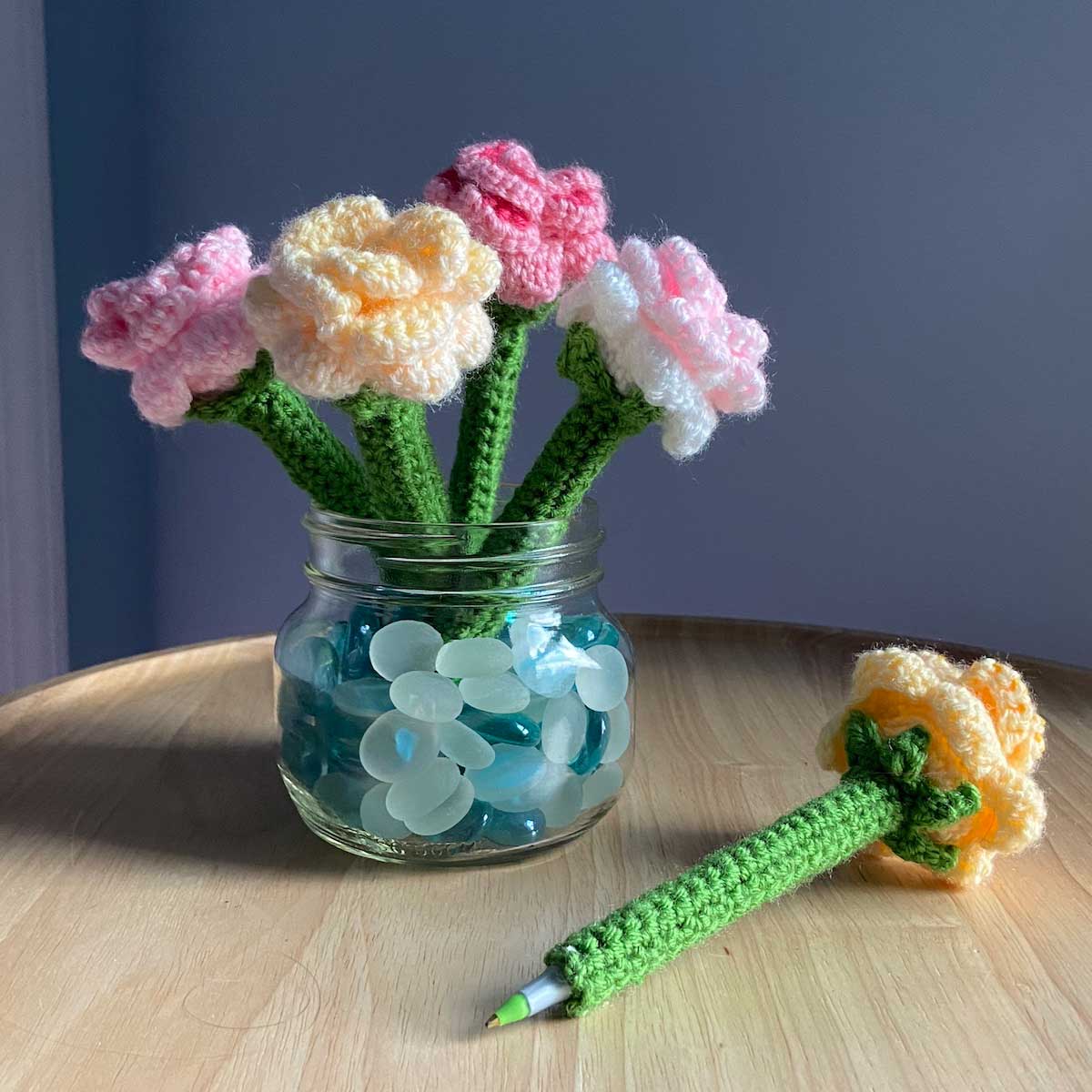 5 crochet flower pens in various colors sit in a glass jar with one sitting out