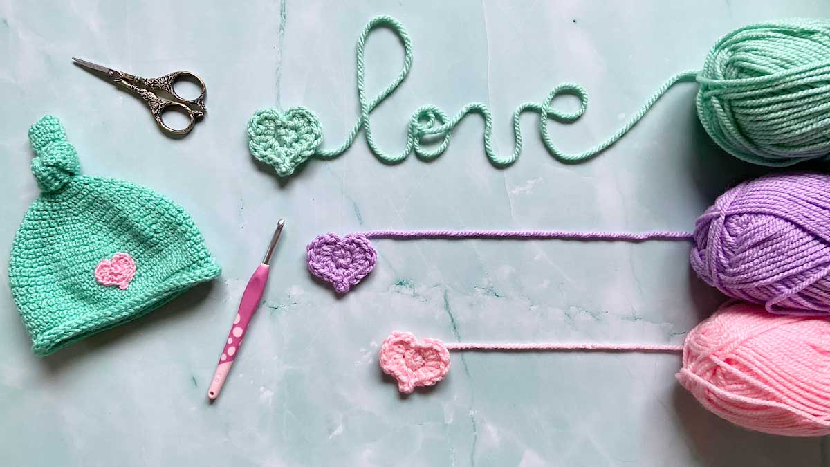 A crocheted baby hat and crocheted hearts lay on a blue background