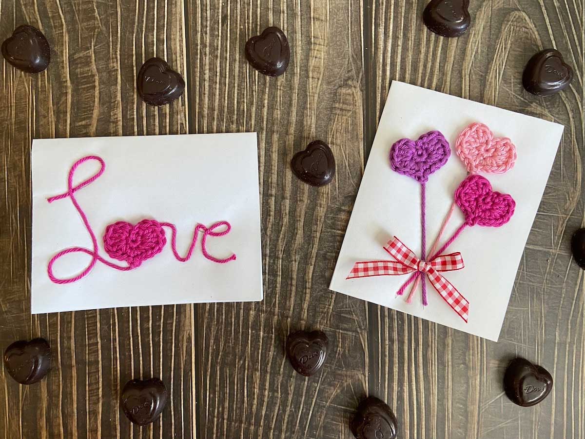 two crocheted valentines day cards lay on wood paneling with chocolates