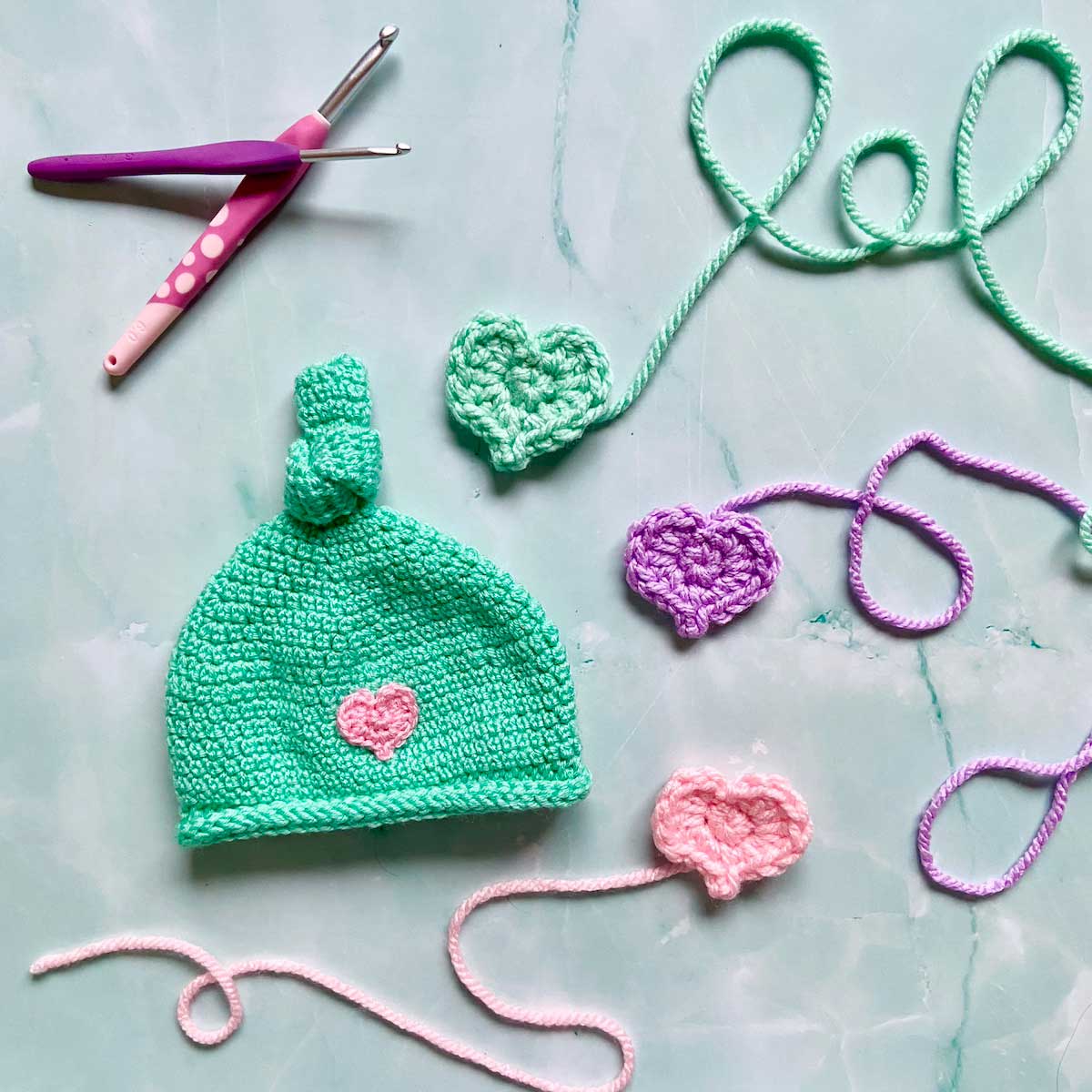 A crocheted baby hat with crocheted hearts in various colors displayed with yarn and crochet hooks