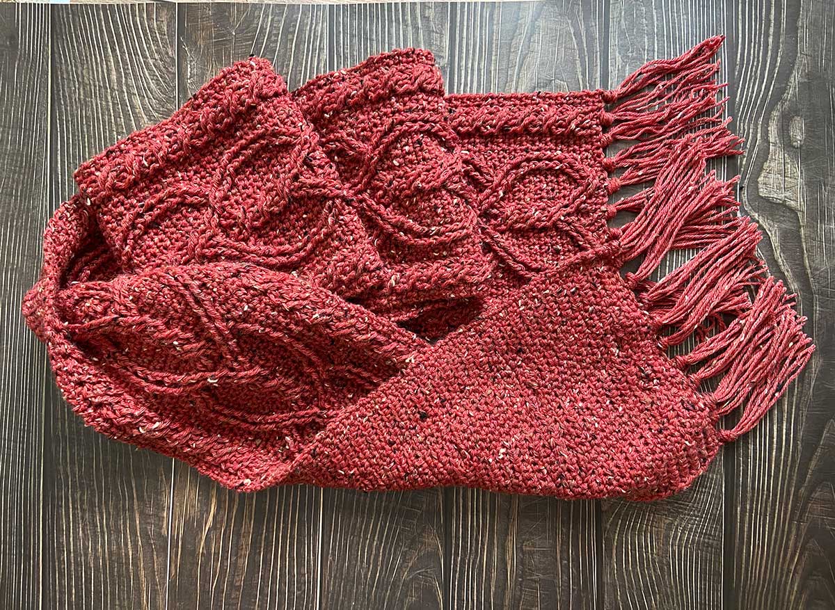 a red crochet cable scarf with a heart motif and fringe lays on a table