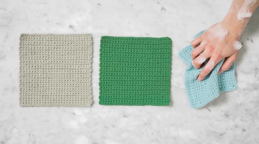 three crocheted dishcloths in tan, green and light blue. The light blue cloth is scrunched up in a hand, covered in suds.