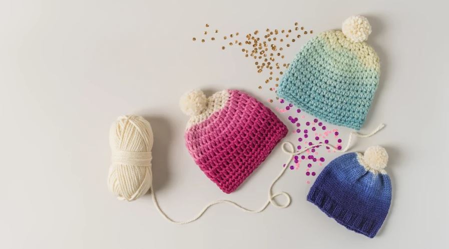 Pink, light blue, and dark blue ombre hats with pompoms on a white background with a skein of white yarn and confetti surrounding the hats.