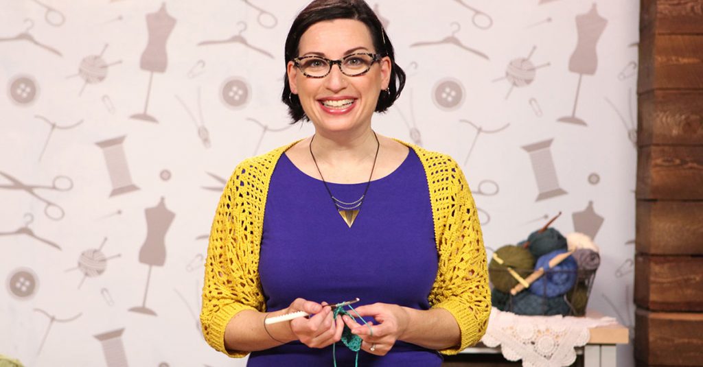 Woman with a purple shirt smiling at the camera and knitting
