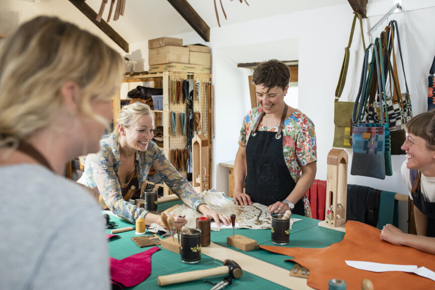 An all-women group undertaking a leather product-making workshop together, they are all standing at the workbench with work tools, ready to make shoes. They are looking at the workshop teacher, waiting for her instruction to start making. / Female Focus Collection