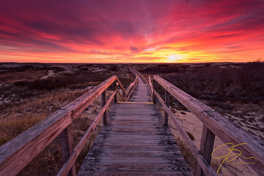 A weathered boardwalk winds through the sand dunes towards the ocean and a fiery sunrise.