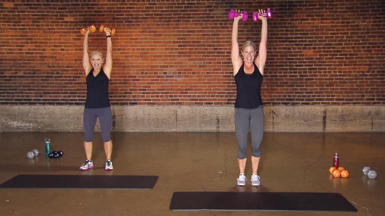 Two women doing dumbbell presses in front of a brick wall