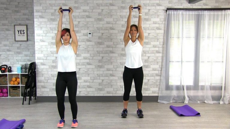 Two women wearing white tank tops workout with dumbbells overhead