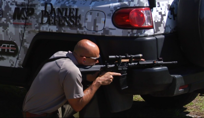 Man aiming a gun around the bumper of a PDN vehicle