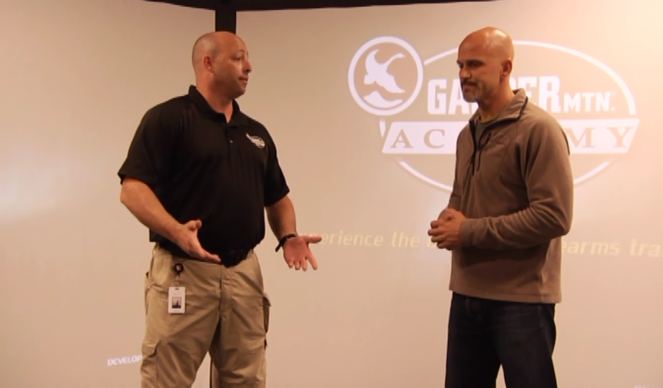 Two men talking in front of a screen with the Gander Mountain Academy logo