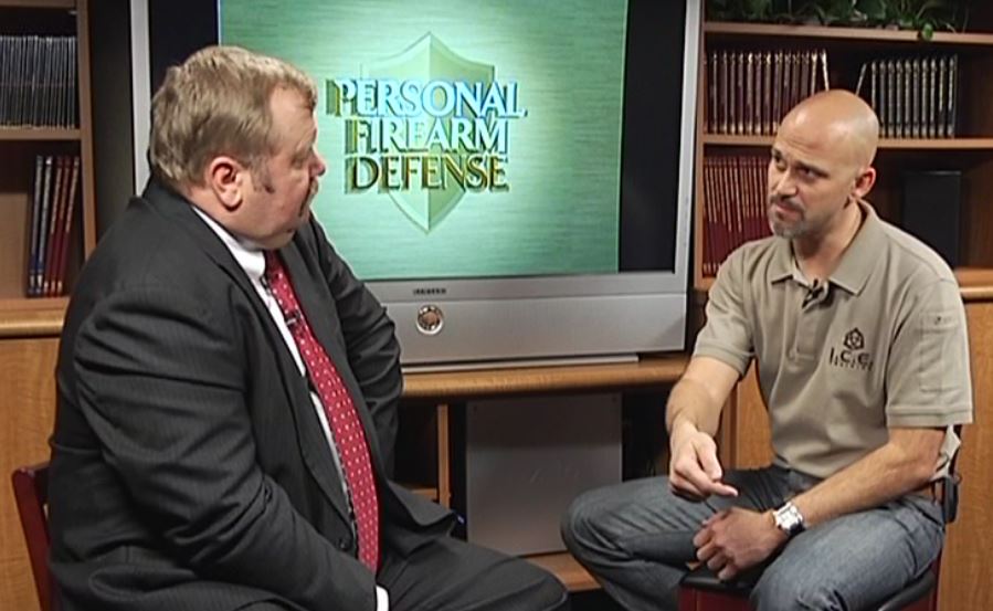 Two men talking by a TV that says personal defense network
