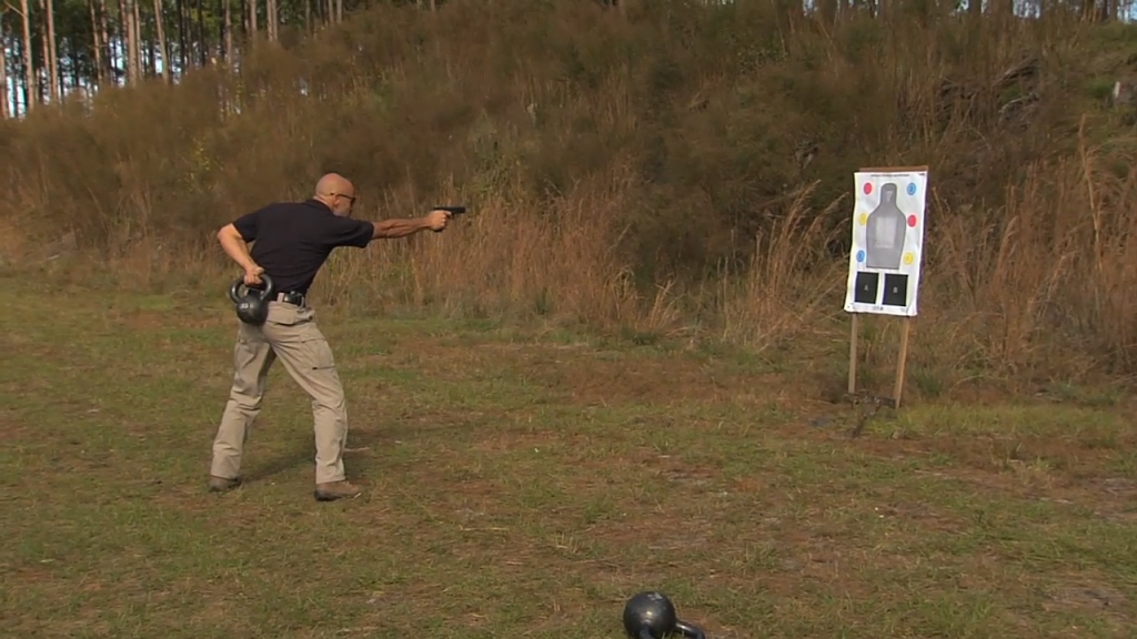 Man aiming a gun while holding a kettlebell behind his back
