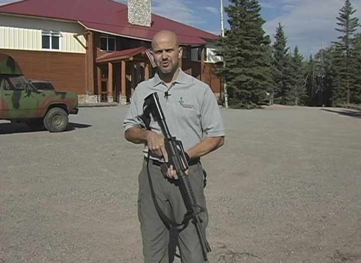 Man outside a building holding a long gun pointed at the ground