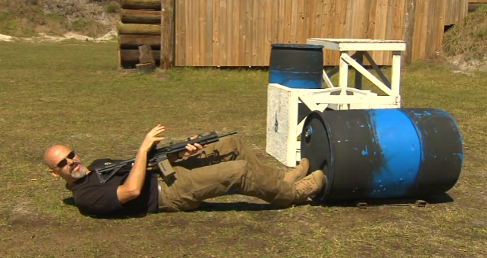 Man on his back with an AR-15 near a barrel