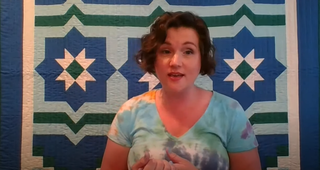 Screenshot of a woman talking in front of a quilt