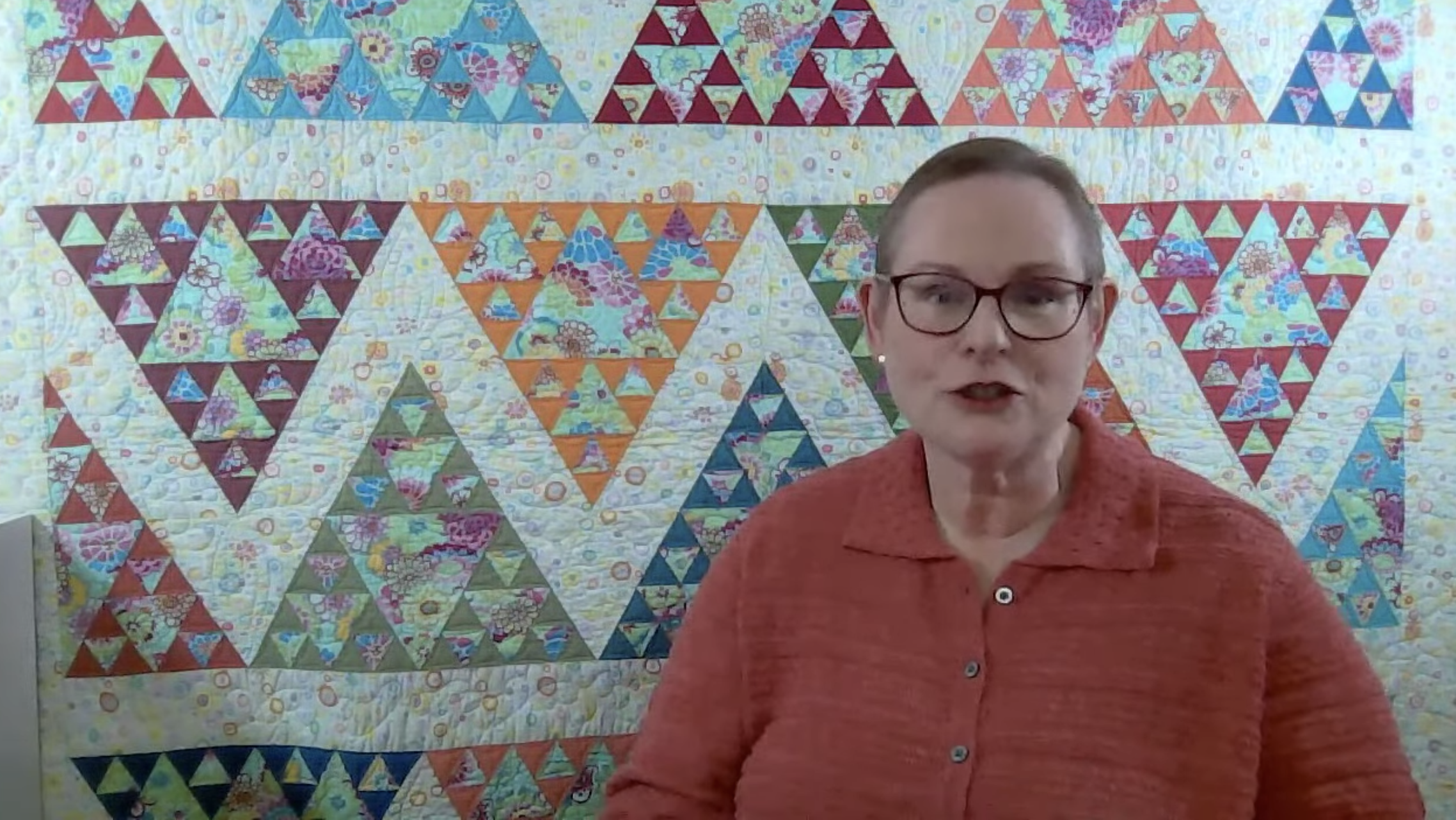 Screenshot of a woman talking in front of a quilt